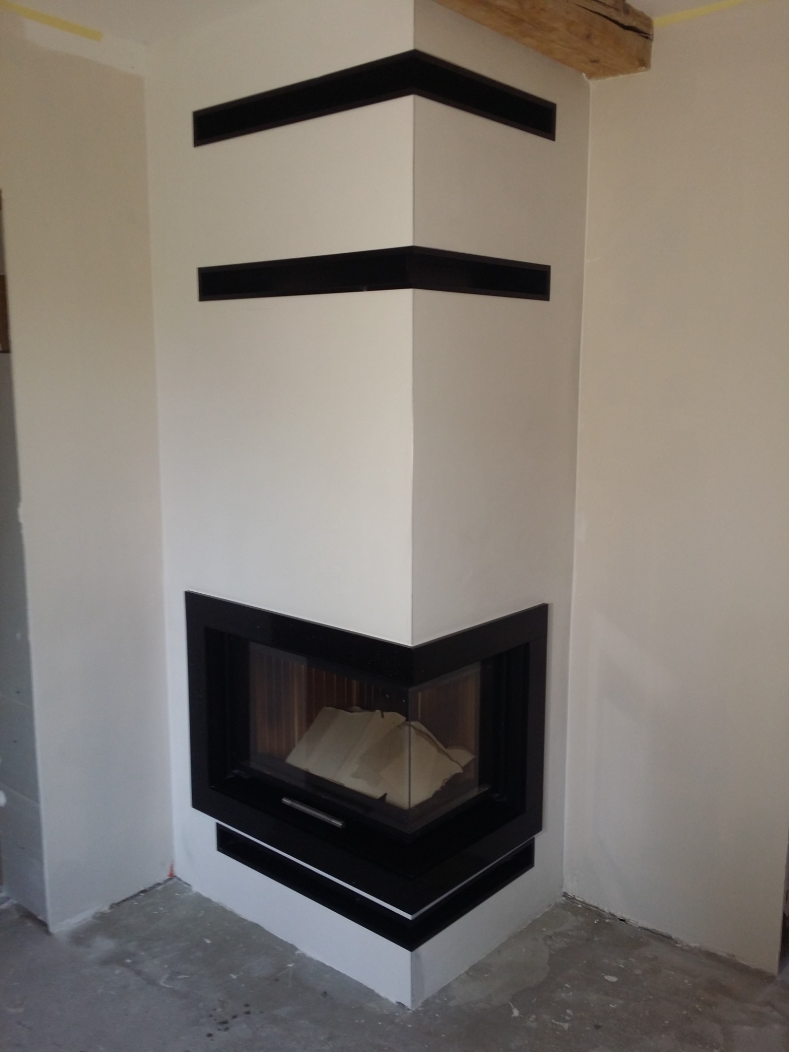 Nowoczesny narożny kominek z czarną obudową w trakcie montażu, widoczne surowe ściany i niedokończona podłoga.