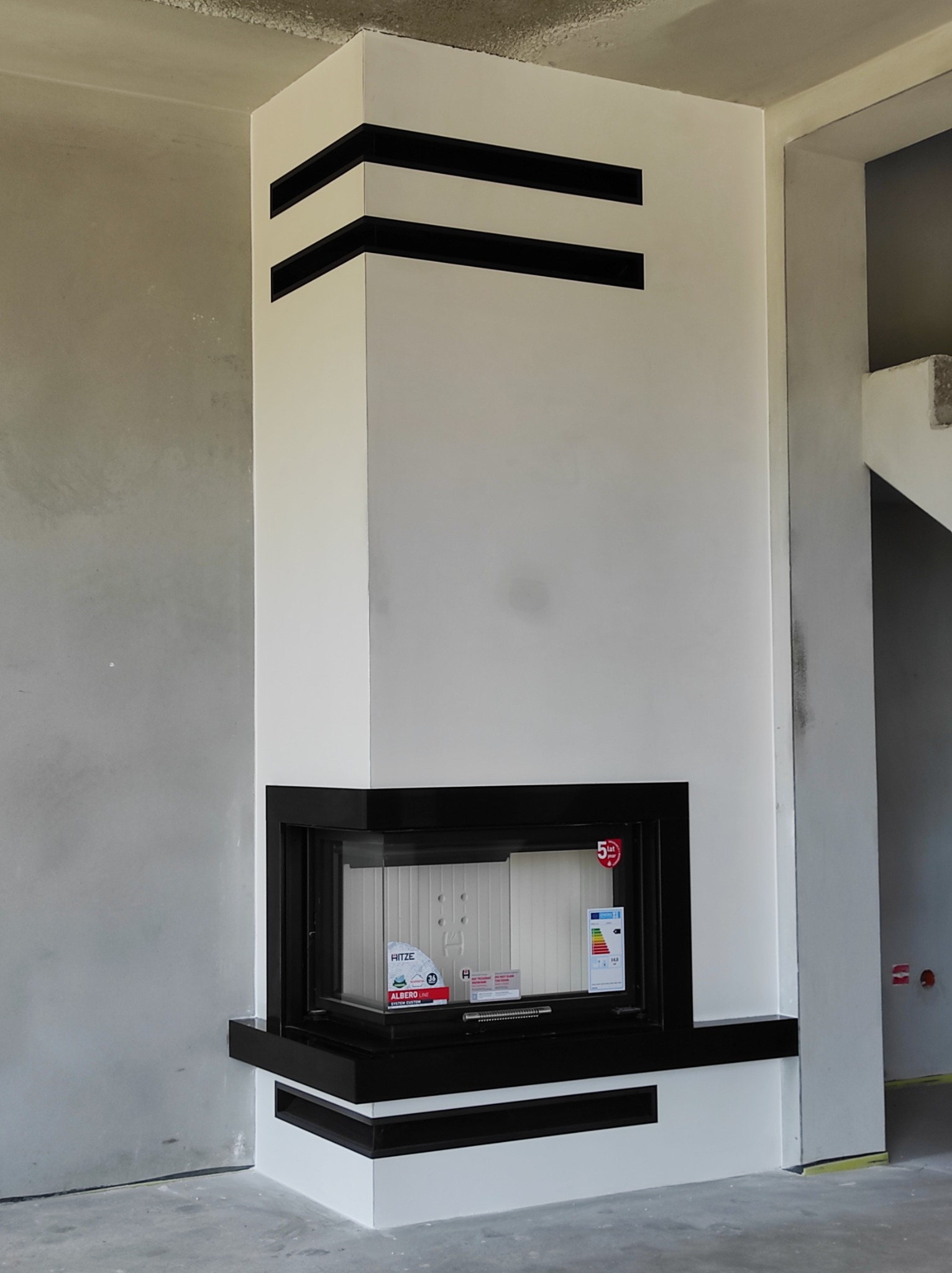 Nowoczesny kominek narożny z białej zabudowy z czarnymi akcentami, widoczny w surowym wnętrzu w trakcie wykańczania.