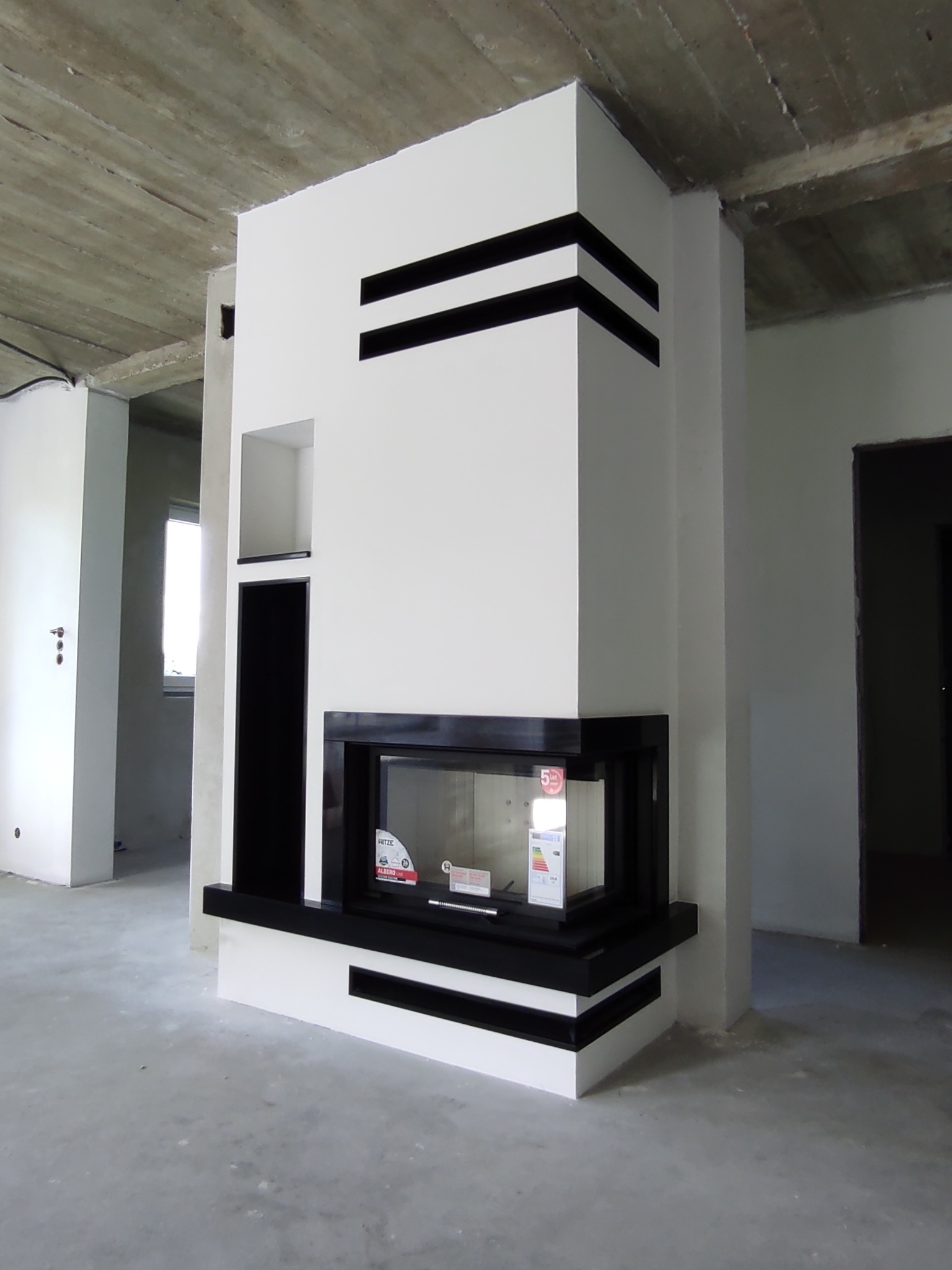 Nowoczesny, biały piec z czarnymi akcentami w surowym wnętrzu, widoczne elementy konstrukcyjne sufitu i niezakończone wykończenie ścian.