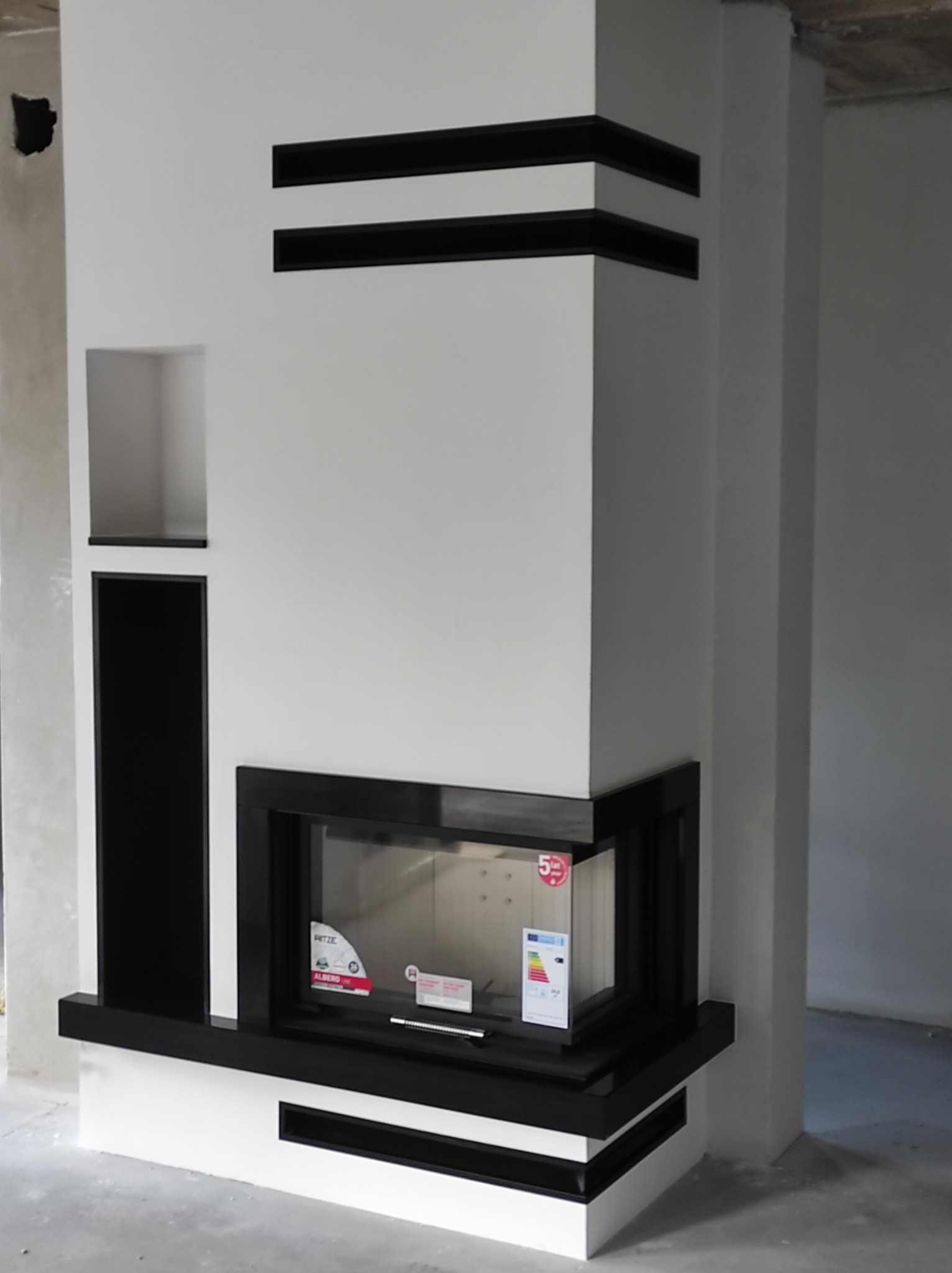 Nowoczesny wkład kominkowy z czarnymi akcentami w obudowie z wnęką, widoczne naklejki informacyjne na szybie, surowy stan wykończenia wnętrza.