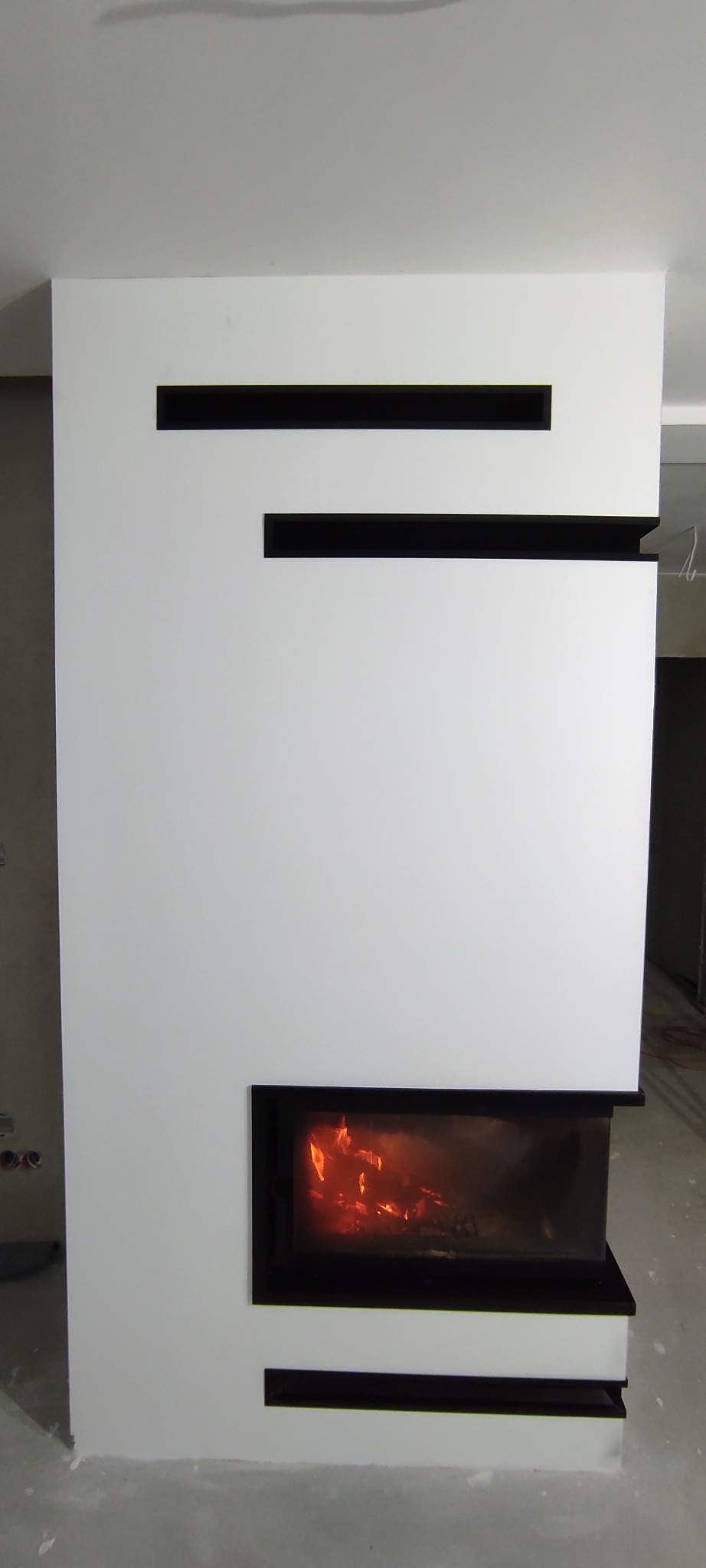 Nowoczesny, biały obudowany wkład kominkowy z czarnymi, poziomymi wnękami i widocznym płomieniem.