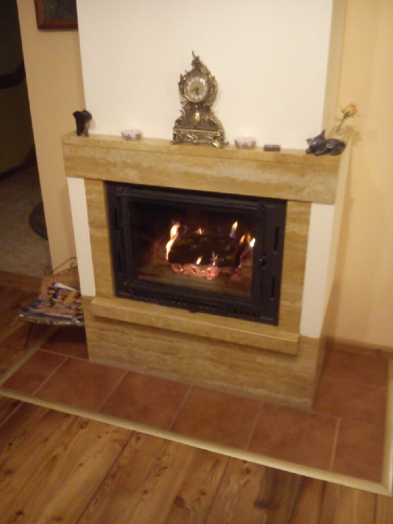 Wnętrze salonu z obudowanym w jasny kamień paleniskiem, na którym stoi ozdobny zegar i drobne dekoracje. W palenisku widoczne płomienie ognia.