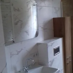 Nowoczesna łazienka z umywalką nablatową na drewnianej szafce, marmurowe płytki na ścianach i lustro z oświetleniem LED.