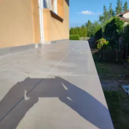 Wyłożony jasnoszarymi płytkami taras przy domu z beżową elewacją, cień osoby robiącej zdjęcie widoczny na płytkach, ogród z zielenią w tle.