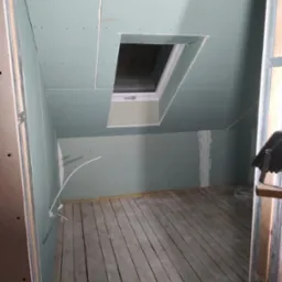 Poddasze z oknem dachowym w trakcie wykańczania płytami gipsowo-kartonowymi, widoczne łączenia płyt wymagające szpachlowania, surowa drewniana podłoga.