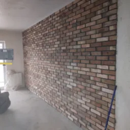 Ściana wykończona cegłą klinkierową w surowym wnętrzu, widoczne niedoskonałości i przewody elektryczne, lampa budowlana w tle.