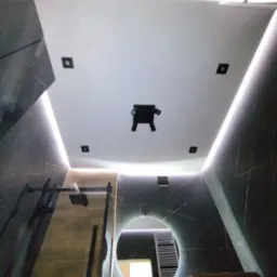 Nowoczesna łazienka z podświetlanym sufitem napinanym, czarne marmurowe ściany z żyłkowaniem, okrągłe lustro z podświetleniem LED i widoczna kabina prysznicowa.