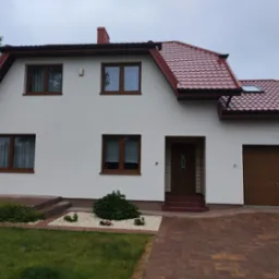 Dom jednorodzinny z czerwoną dach&oacute;wką, brązowymi oknami i garażem, otoczony zielonym trawnikiem i ozdobnymi krzewami.