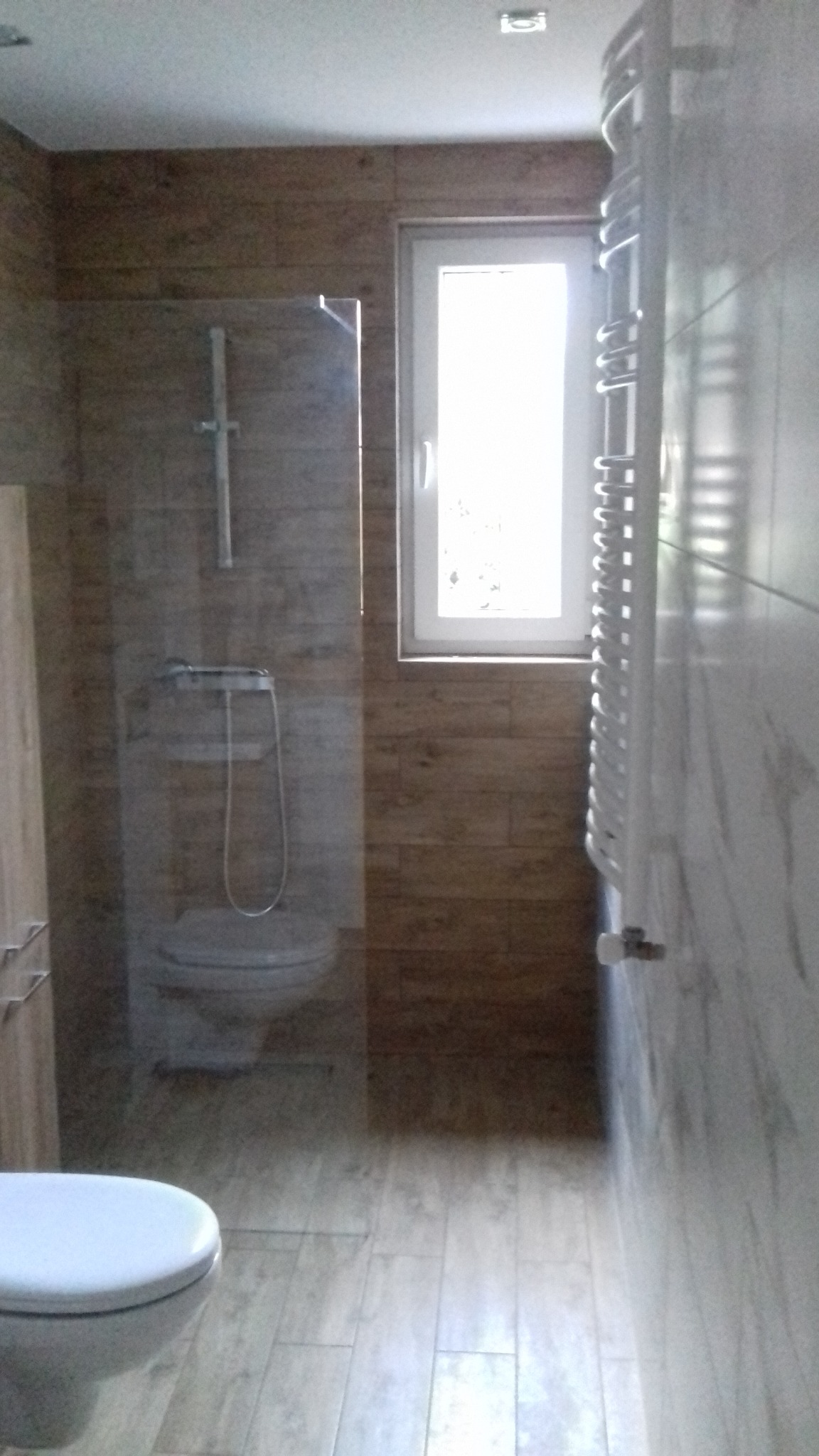 Wykończona łazienka z kabiną prysznicową za szkłem, drewnopodobnymi płytkami na ścianach i podłodze, białym grzejnikiem drabinkowym oraz oknem z białymi ramami.