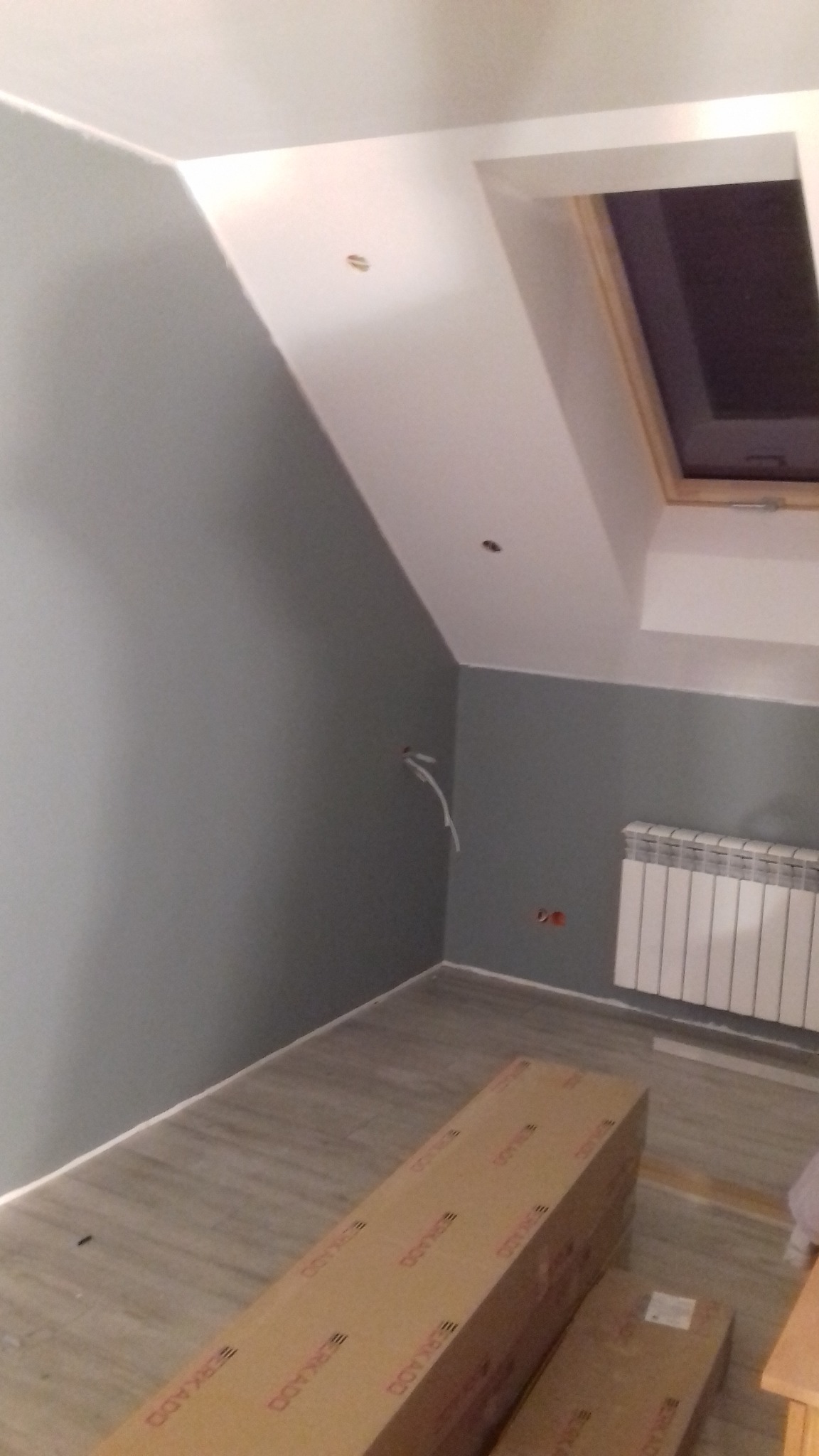 Poddasze po częściowym remoncie, szare ściany, drewniane panele podłogowe, okno dachowe, grzejnik, kartony z panelami podłogowymi, widoczne kable wystające ze ściany.