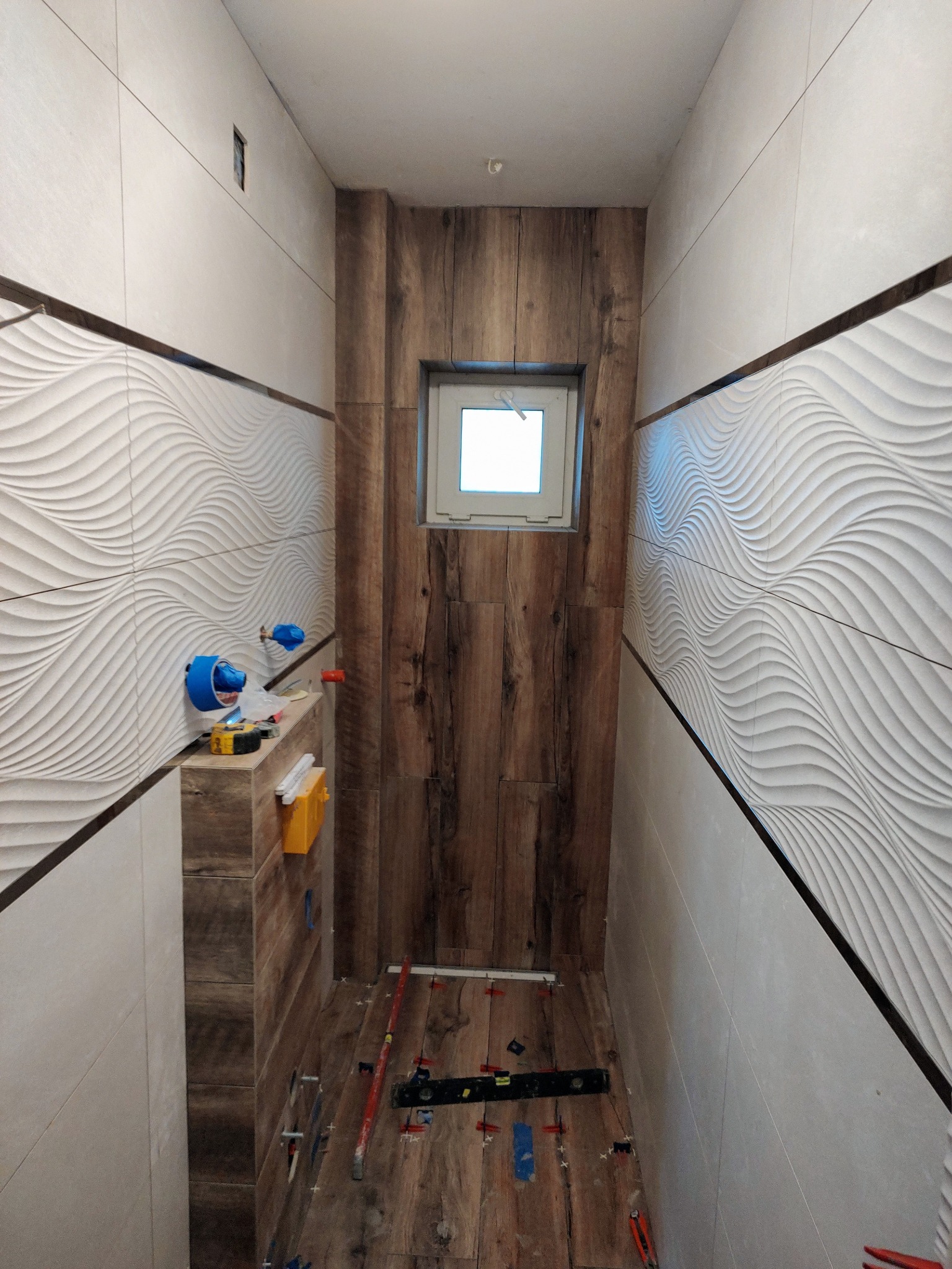 Wąska łazienka w trakcie wykańczania: ułożone płytki imitujące drewno na podłodze i ścianie z oknem, obok ściana z białych płytek 3D z poziomym wzorem fal, widoczne narzędzia glazurnicze i poziomica.