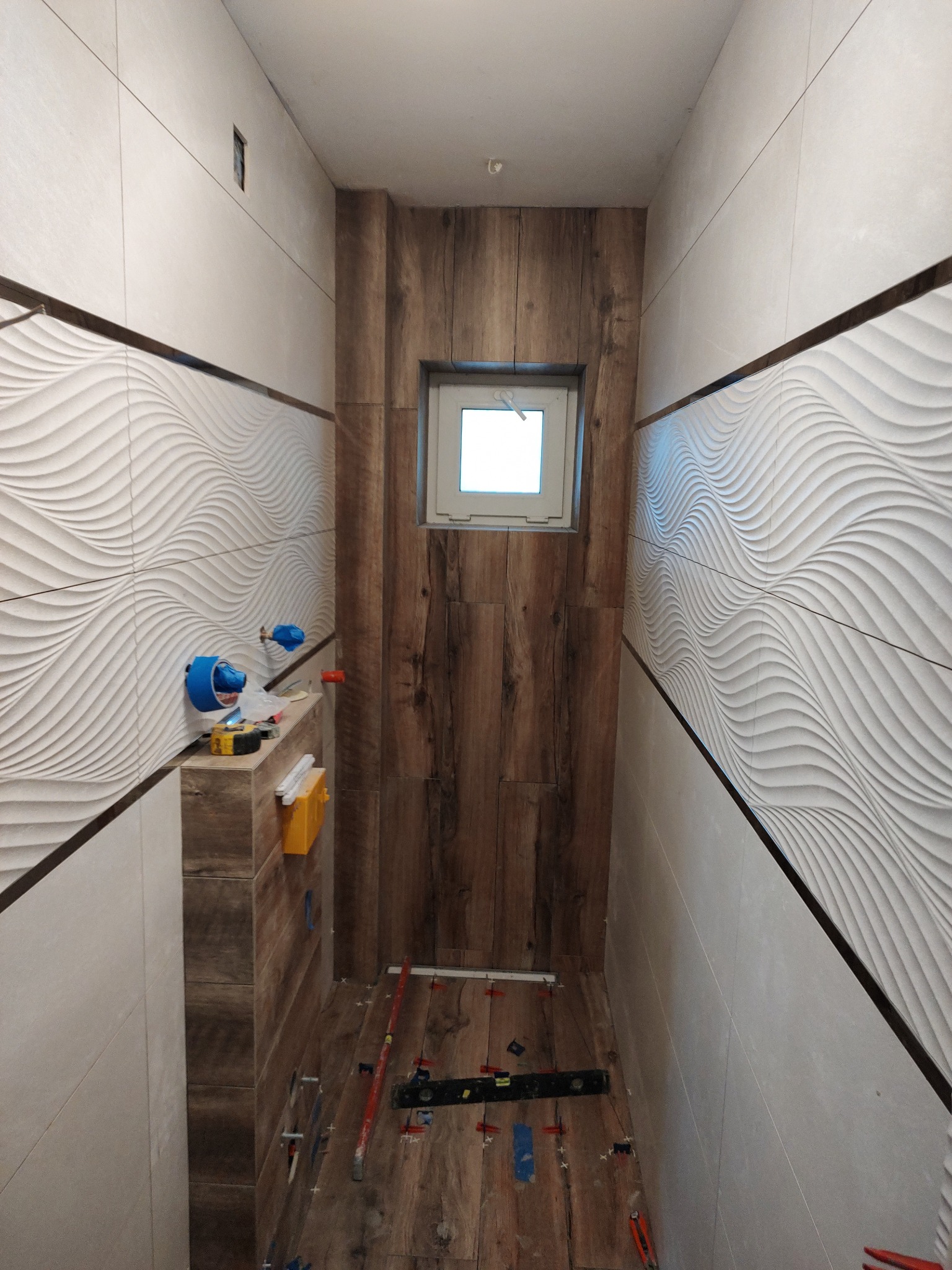Wąska łazienka w trakcie wykańczania, z ułożonymi płytkami imitującymi drewno na podłodze i części ściany, oraz falistymi, białymi płytkami na pozostałej części ścian. Widoczne narzędzia...