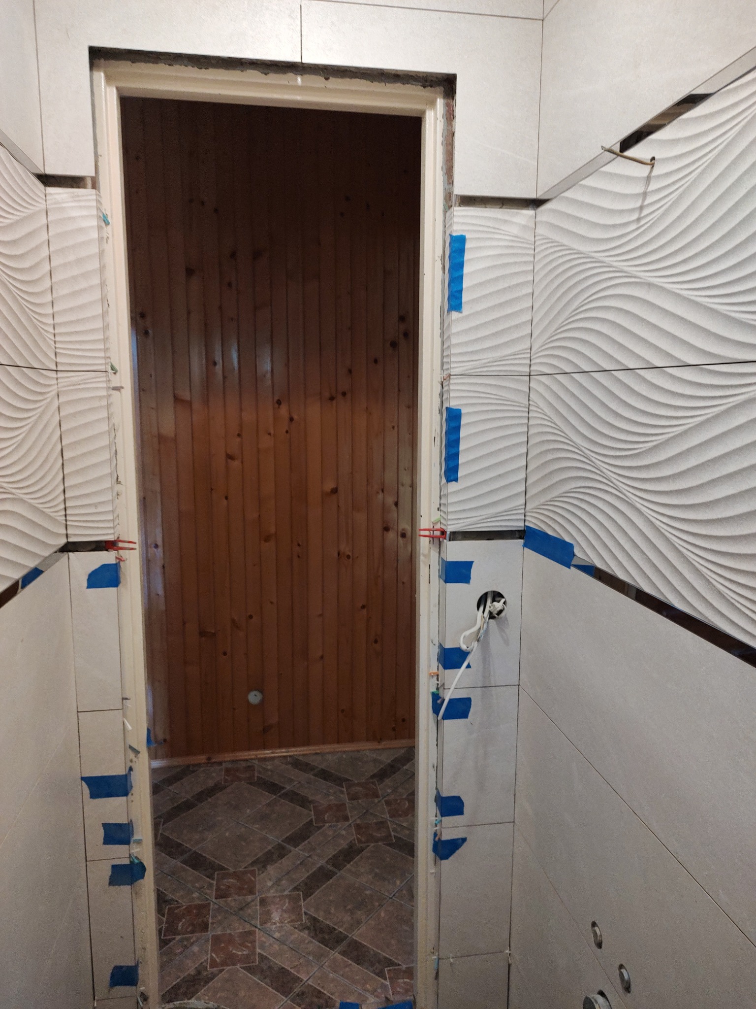 Łazienka w trakcie remontu: widoczne płytki ścienne z falistym wzorem, zabezpieczone niebieską taśmą, oraz fragment drewnianej ściany i podłogi w przejściu.