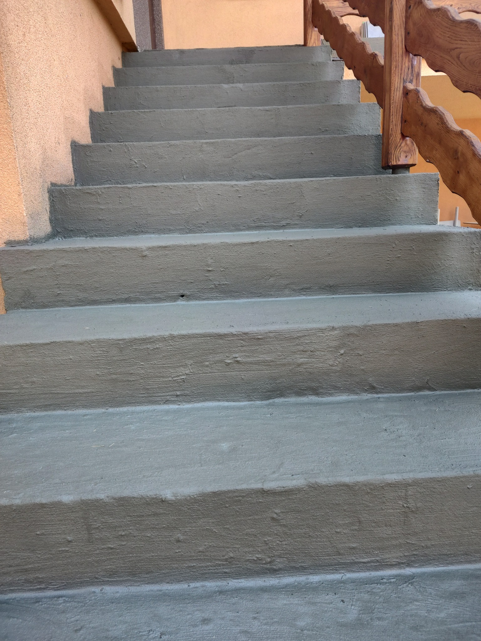 Wykończone betonowe schody zewnętrzne z drewnianą balustradą o falistym wzorze, pomalowane na szaro, widoczne z dołu.