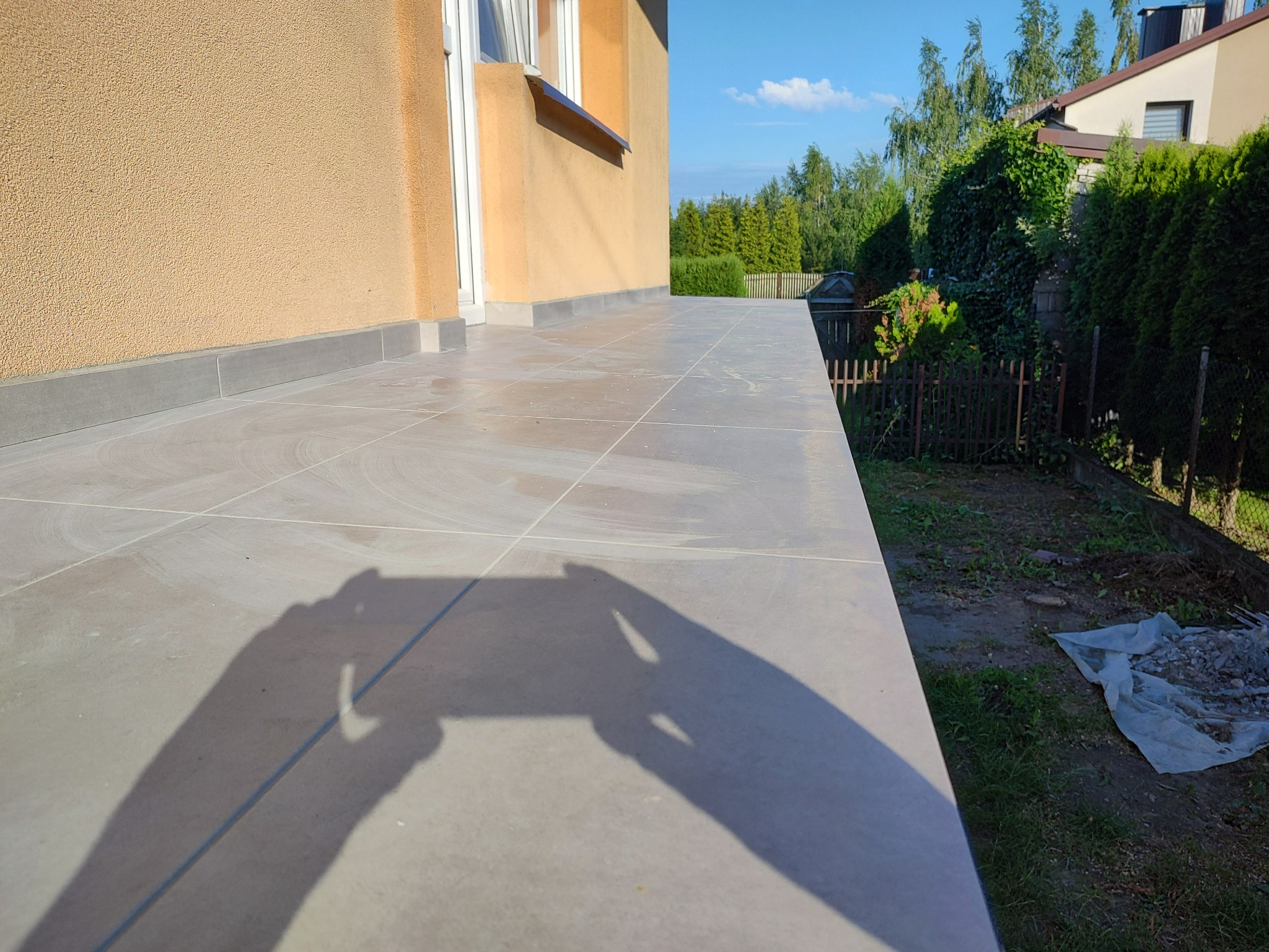 Wyłożony jasnoszarymi płytkami taras przy domu z beżową elewacją, cień osoby robiącej zdjęcie widoczny na płytkach, ogród z zielenią w tle.