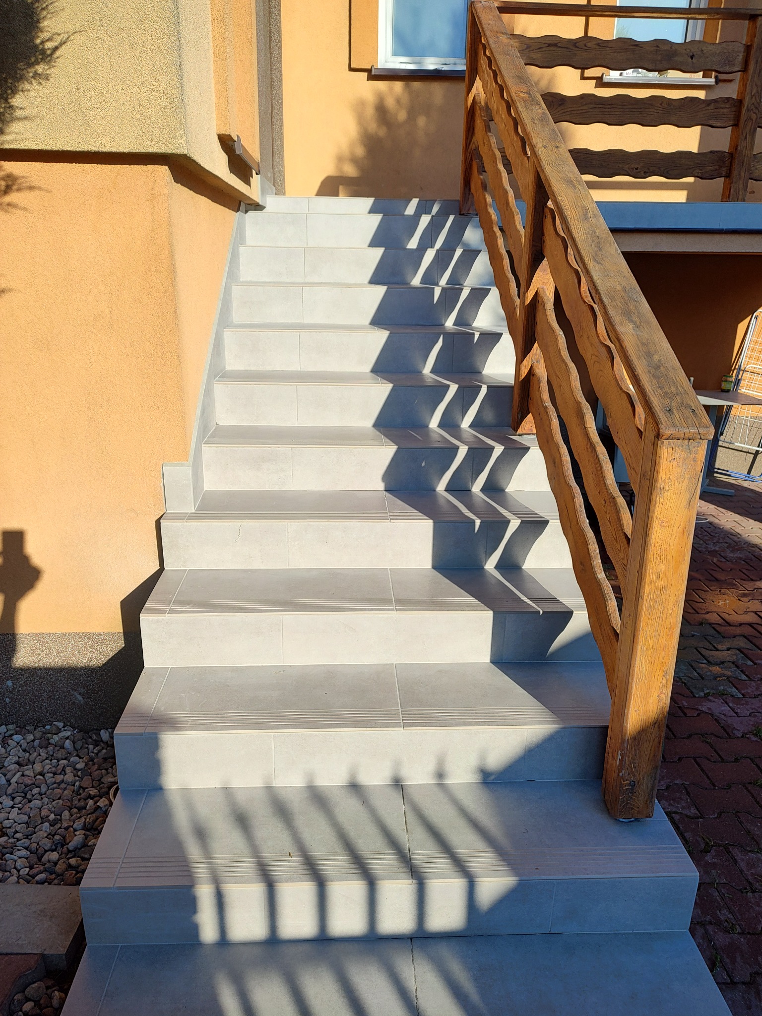 Zewnętrzne schody wyłożone szarą terakotą z ozdobnymi paskami antypoślizgowymi, z drewnianą balustradą o falistym wzorze, oświetlone popołudniowym słońcem rzucającym cienie na stopnie.
