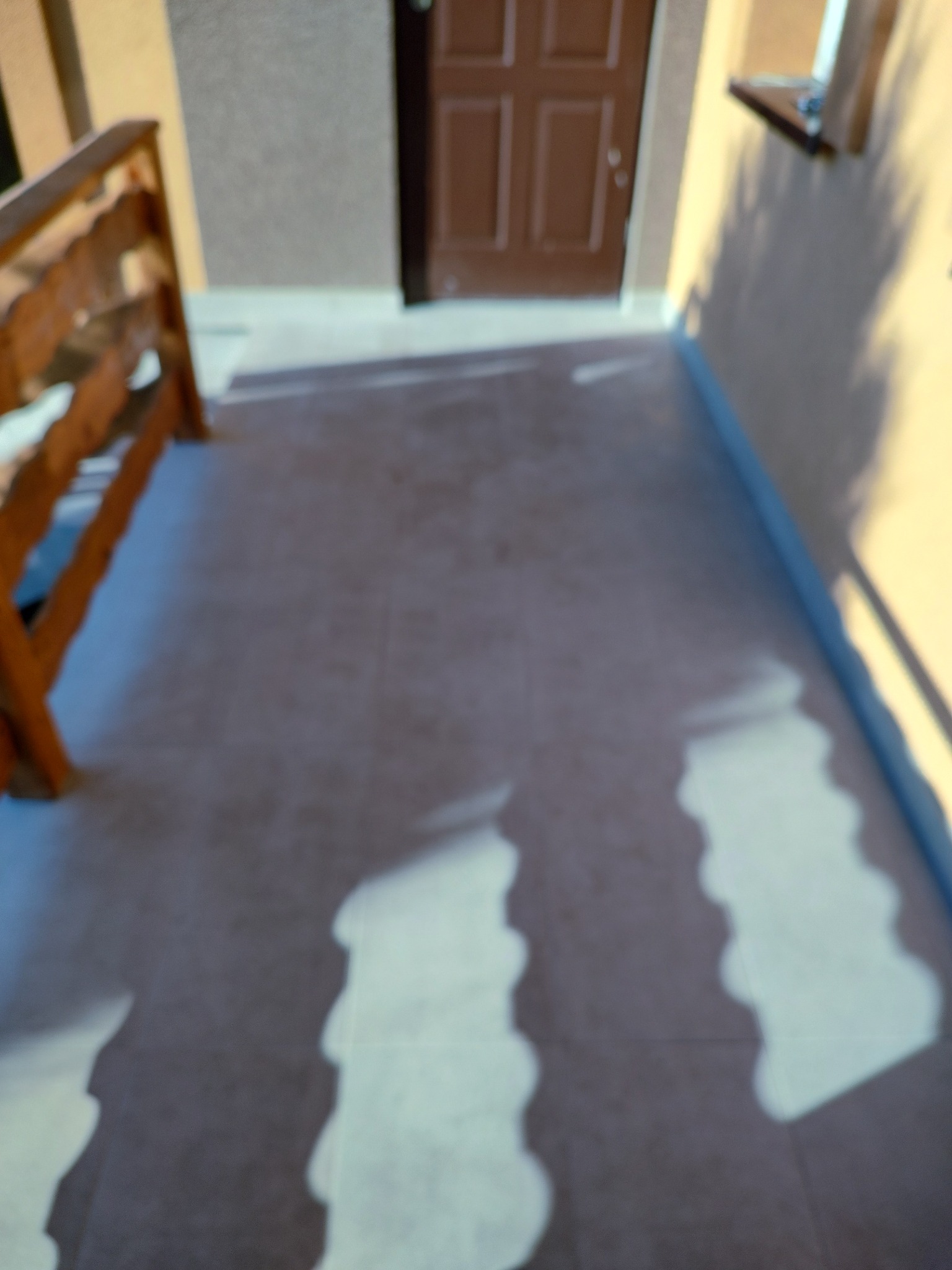 Wyłożony płytkami taras przed wejściem do domu, z widoczną drewnianą barierką i cieniami drzew.