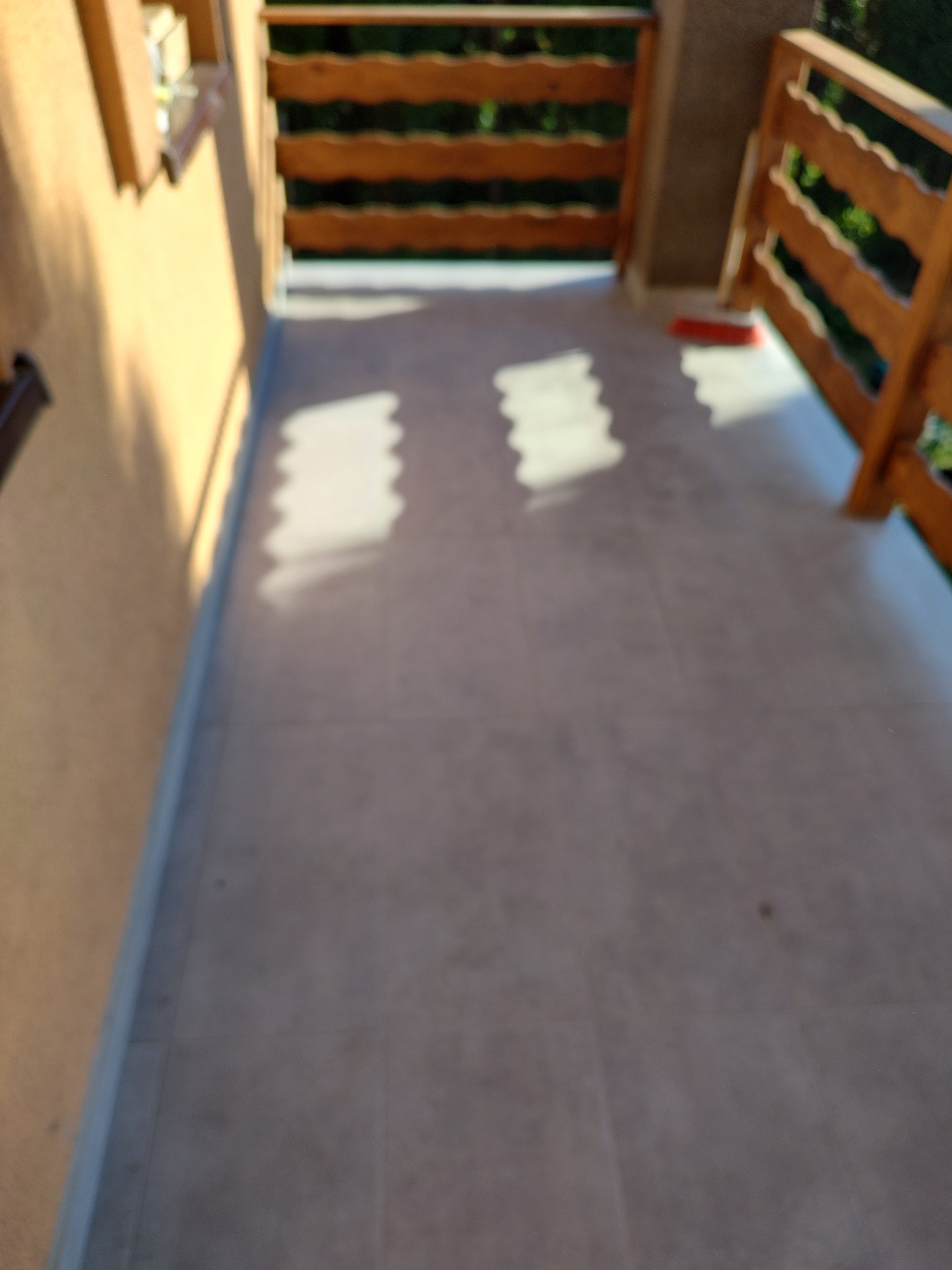 Świeżo wyłożona, jasnoszara podłoga balkonu z drewnianą balustradą, oświetlona popołudniowym słońcem, tworzącym geometryczne cienie.