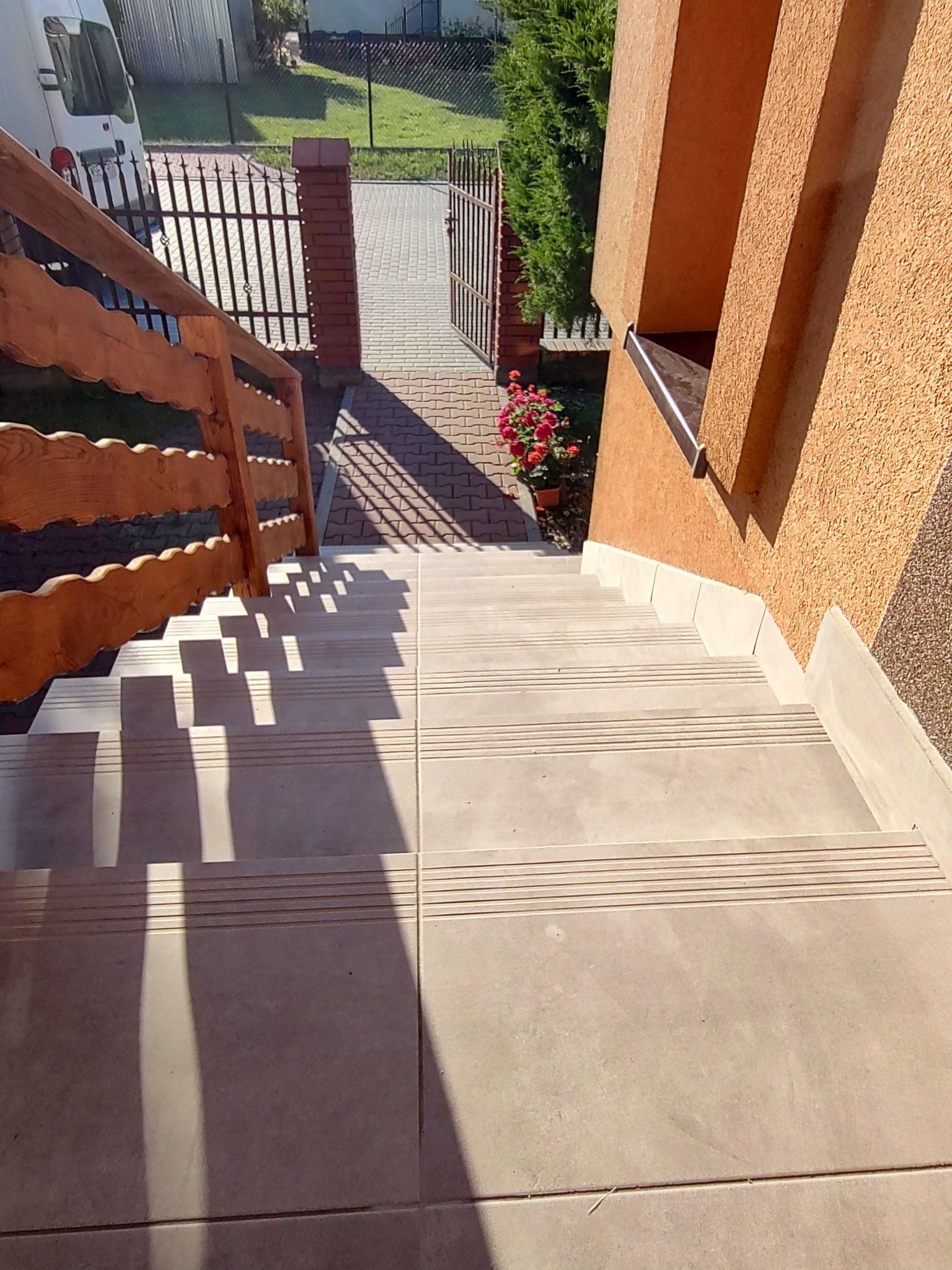 Zewnętrzne schody wyłożone beżowymi płytkami z antypoślizgowymi rowkami, drewniana balustrada z falistym wzorem, elewacja budynku o pomarańczowej strukturze baranka, widok na furtkę i podjazd...