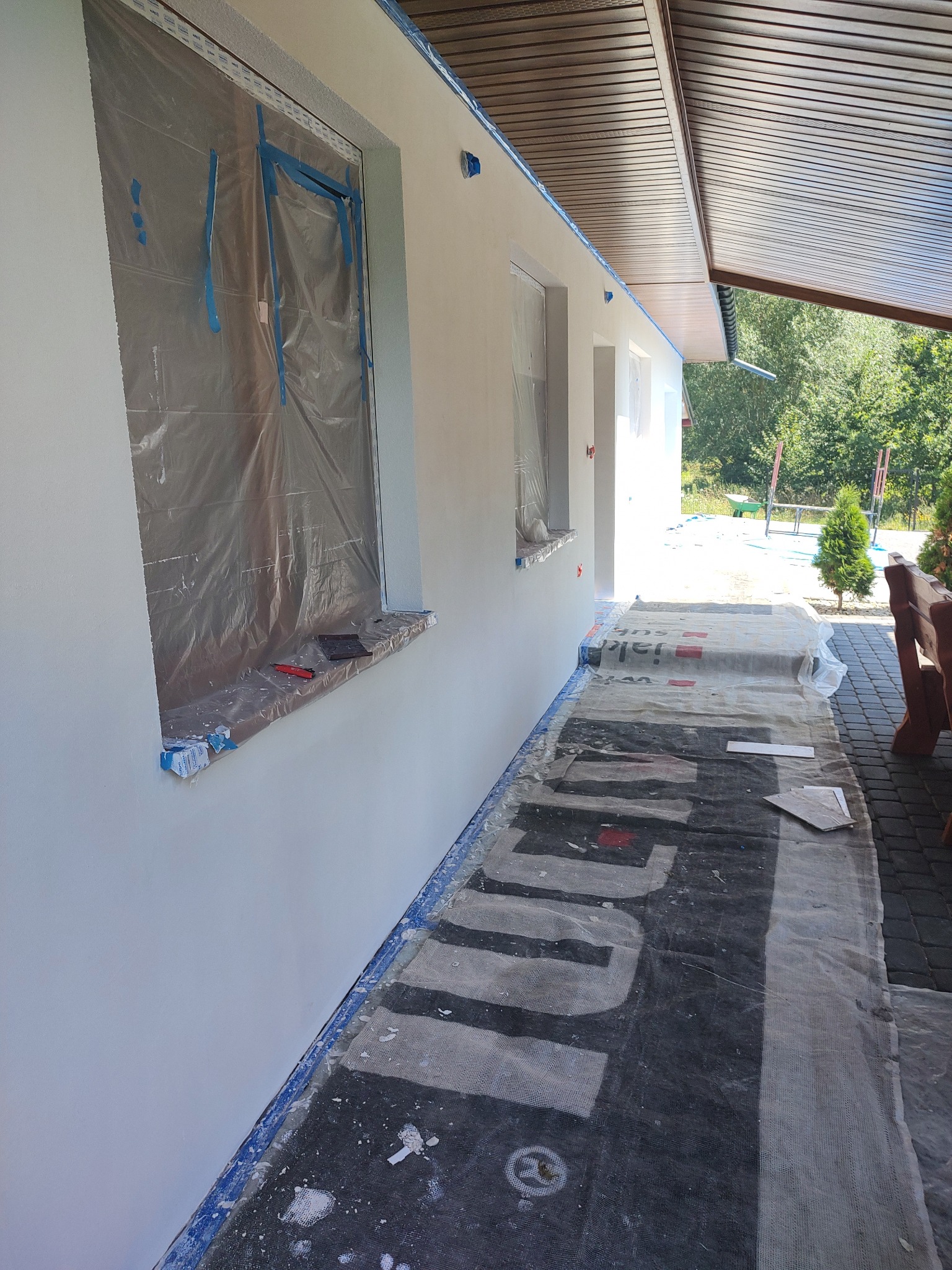 Elewacja domu po świeżym malowaniu, okna zabezpieczone folią i taśmą malarską, podłoże chronione matą, widoczne ślady farby.