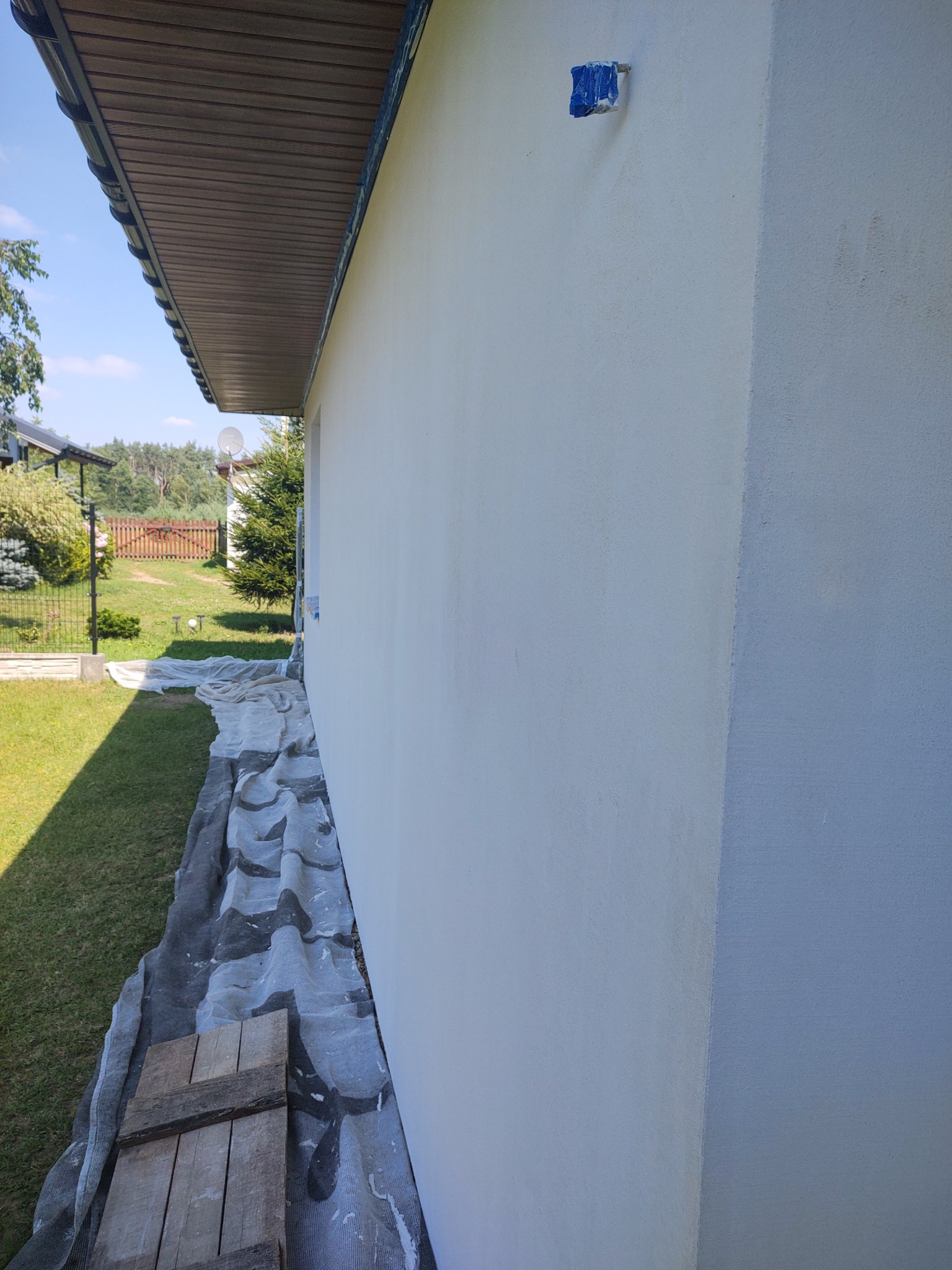 Świeżo otynkowana ściana domu jednorodzinnego, zabezpieczona folią malarską i drewnianymi deskami, z widocznym fragmentem podbitki dachowej i niebieskim zabezpieczeniem na elewacji.