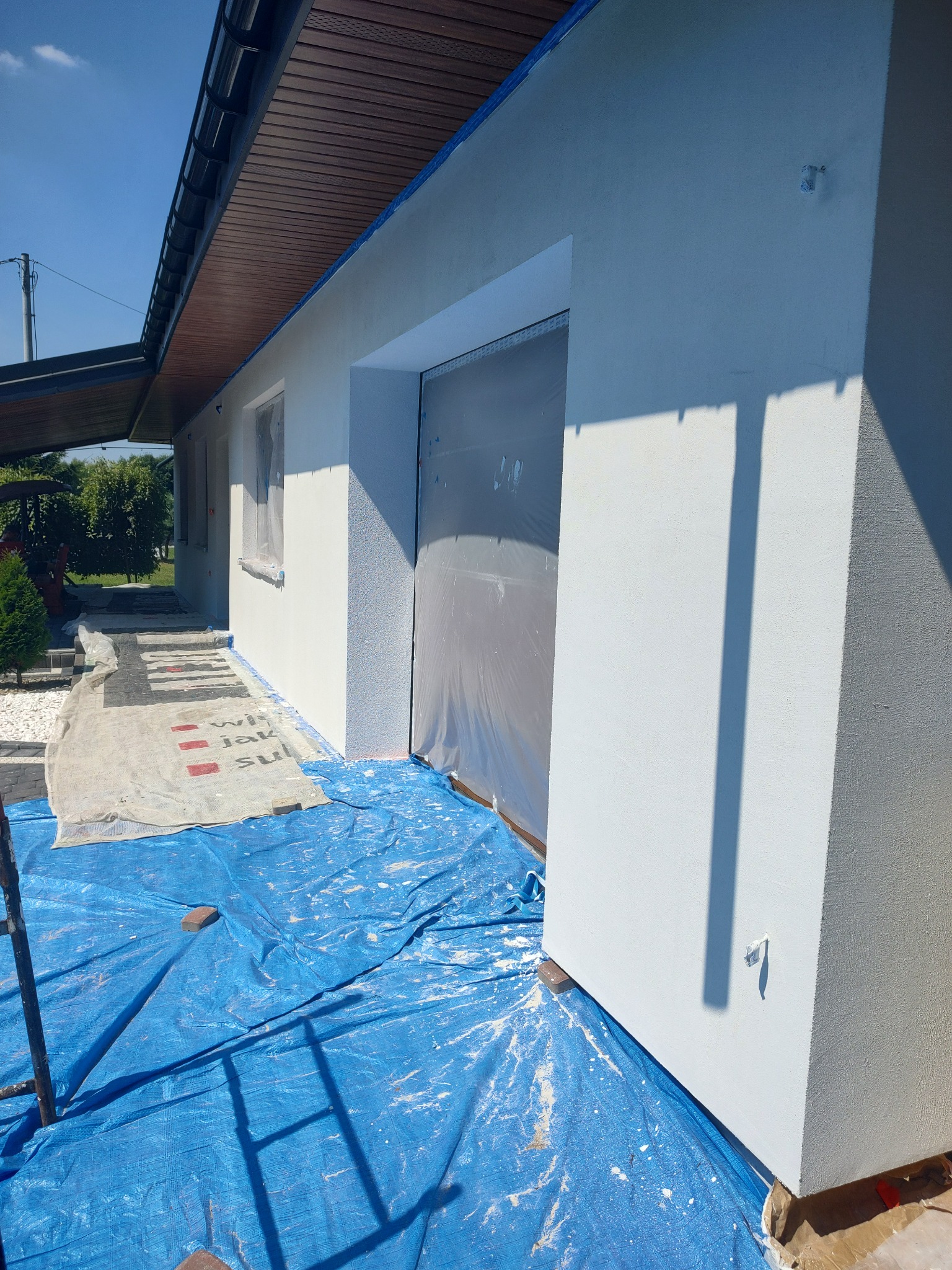 Biała elewacja domu jednorodzinnego w trakcie malowania, zabezpieczona folią i plandekami, widoczny fragment rusztowania i drewniane podbitki dachu.