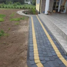 Ułożona ścieżka z kostki brukowej w kolorze grafitowym z żółtymi pasami, prowadząca od trawnika do schodów wejściowych domu, z małym psem siedzącym na schodach.
