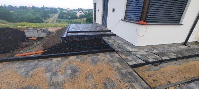 Układanie kostki brukowej w trakcie prac: widoczne ułożone schody i fragment tarasu z kostki, przygotowane podłoże z piaskiem, niezabudowana rabata, paleta kostki brukowej w tle.