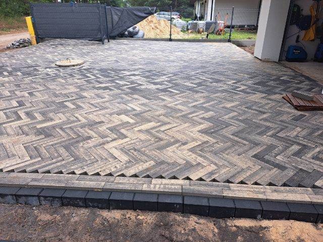 Ułożona dwukolorowa kostka w jodełkę na podjeździe, z widocznym włazem i częściowo wykończonym obrzeżem z ciemnej kostki.