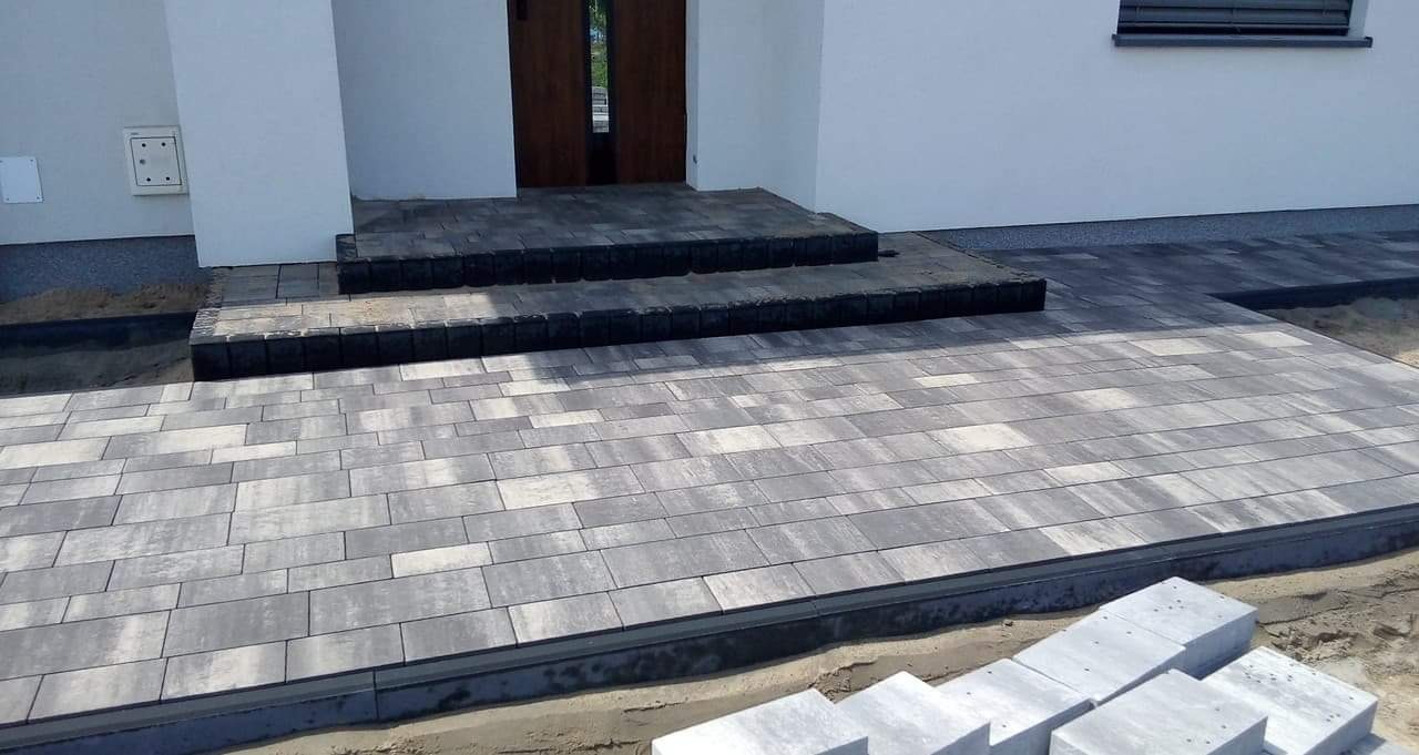 Ułożona kostka brukowa przed domem z dwoma stopniami wejściowymi, widoczne niezakończone prace ziemne i stos kostki obok.