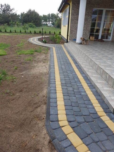 Nowo ułożona ścieżka z ciemnoszarej kostki brukowej z dwoma żółtymi pasami prowadząca do domu z jasną elewacją i małym psem przy kolumnie.