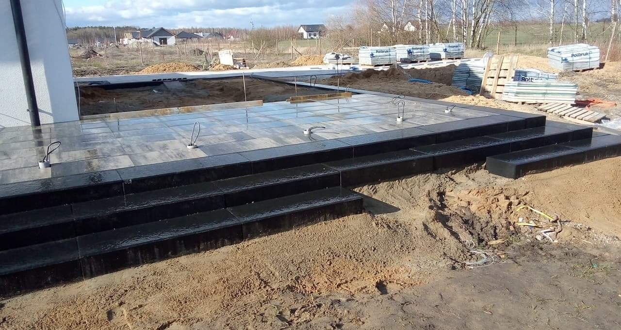 Etap budowy tarasu z czarnej kostki granitowej w Bydgoszczy, widoczne przygotowane podłoże i palety z materiałem w tle.