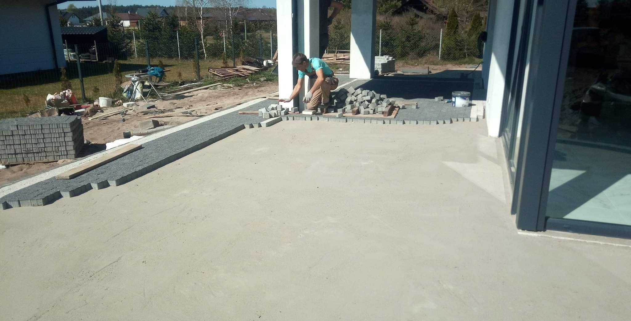 Pracownik układa szarą kostkę granitową wzdłuż fundamentów domu, widoczne stosy kostki i przygotowane podłoże