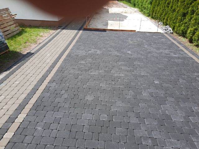 Ukończony podjazd z ciemnoszarej kostki brukowej o regularnym wzorze, obrzeża z jasnej kostki, widoczny fragment budynku i przygotowany teren pod dalsze prace.
