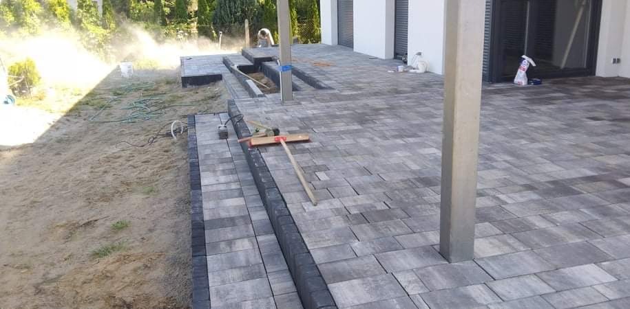 Trwa układanie szarej kostki brukowej na tarasie, widoczne narzędzia i częściowo ułożona powierzchnia. W tle rozpylona woda i prace wykończeniowe.