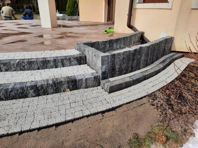 Schody zewnętrzne i rabata z kostki granitowej, jasnoszara kostka ułożona w łuk, widoczne osoby w tle.