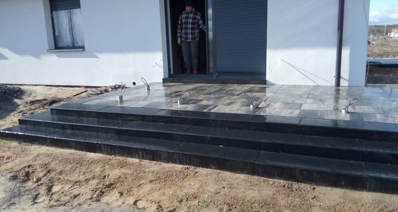 Nowoczesny taras z szarej kostki z trzema stopniami, w tle biały dom z zamkniętymi roletami i mężczyzna w drzwiach.