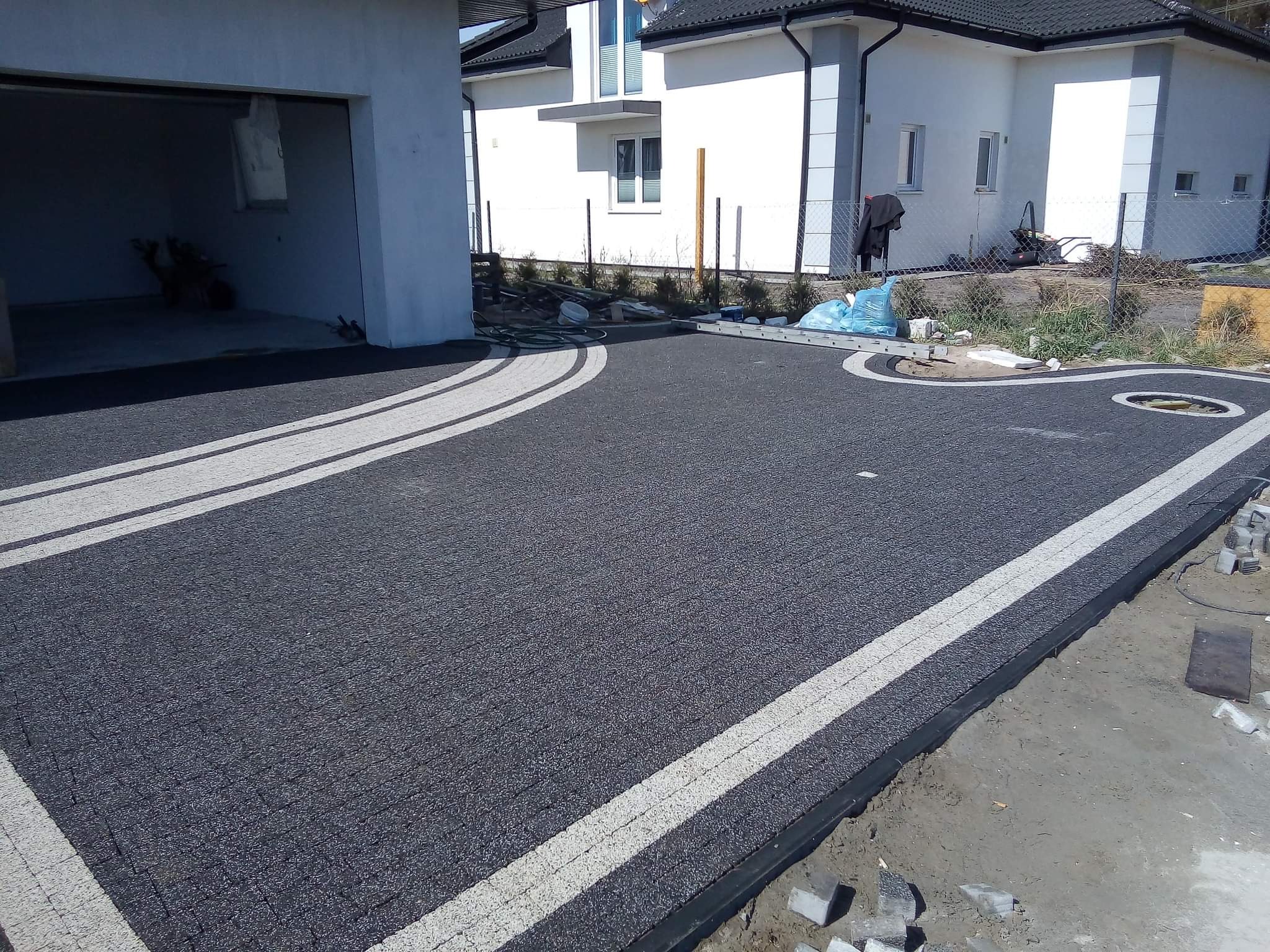 Ułożona nawierzchnia z ciemnej kostki z jasnymi pasami oddzielającymi, widoczne niedokończone obrzeża i elementy kostki przygotowane do ułożenia, w tle dom z garażem.
