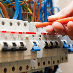 Elektryk w trakcie sprawdzania bezpieczników w skrzynce rozdzielczej przy użyciu próbnika napięcia z pomarańczową rączką.