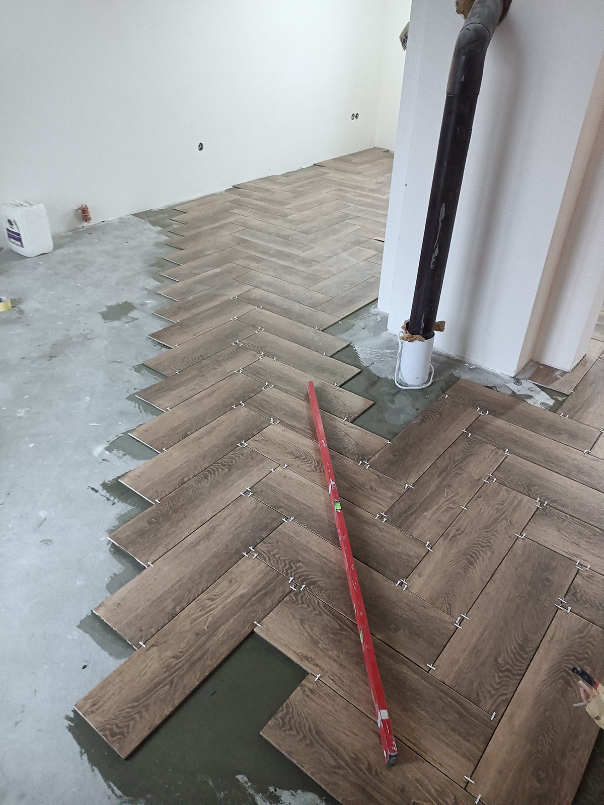 Układanie brązowych płytek podłogowych w jodełkę; widoczny poziomica, krzyżyki dystansowe i fragment surowej podłogi z zaprawą.