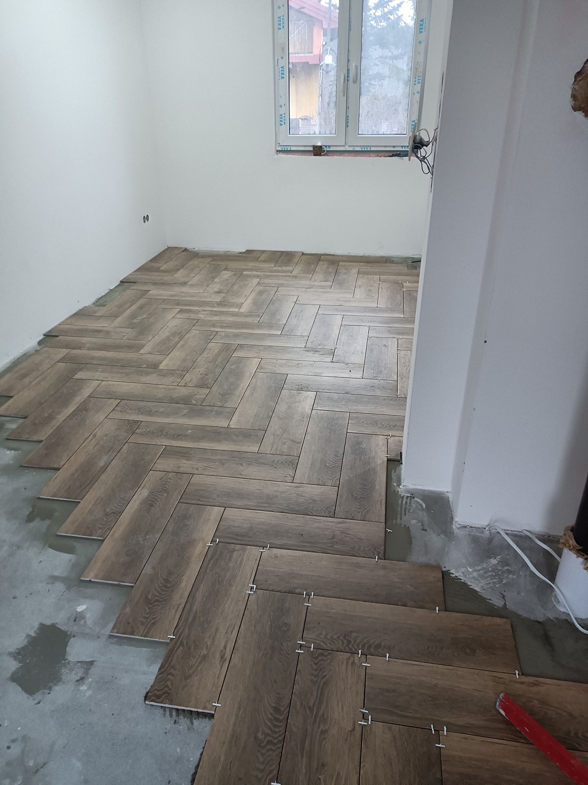 Układanie brązowych płytek podłogowych w jodełkę w jasnym pokoju, widoczne plastikowe krzyżyki dystansowe, niedokończona podłoga.