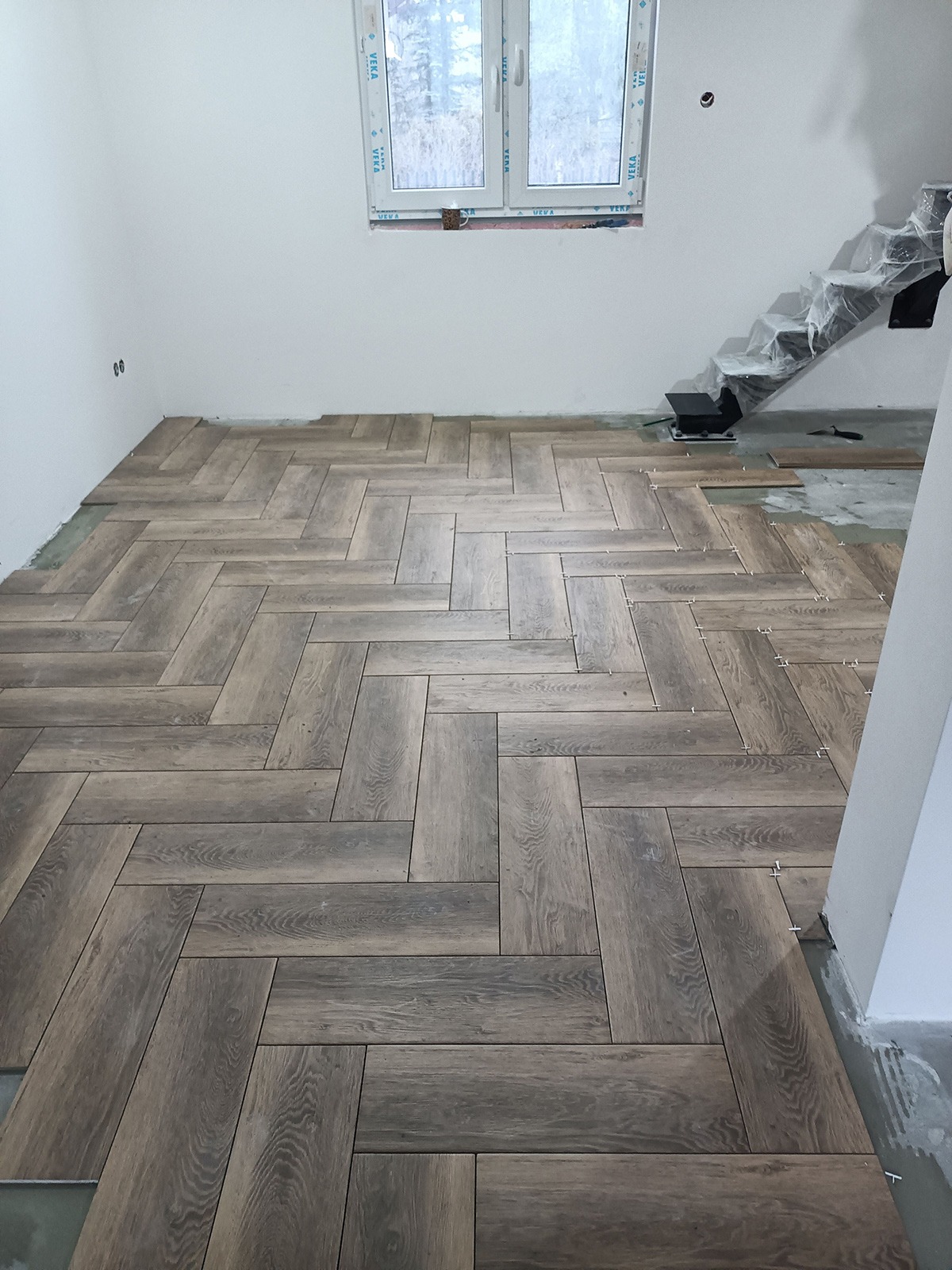 Układanie drewnopodobnych płytek w jodełkę na podłodze w trakcie remontu, widoczne krzyżyki dystansowe i częściowo zakryte folią schody.