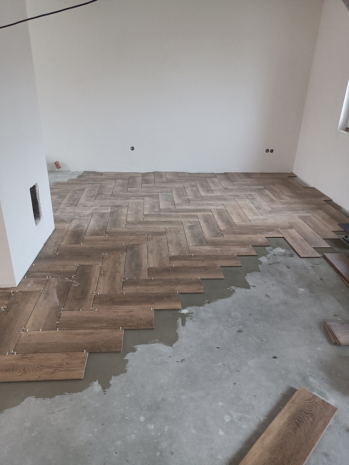 Układanie podłogi z płytek drewnopodobnych w jodełkę, widoczne krzyżyki dystansowe i świeży klej.
