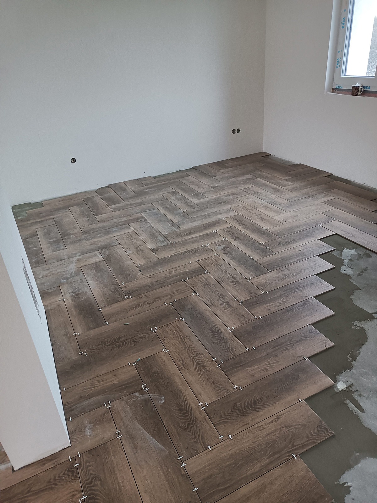 Układanie podłogi z płytek imitujących drewno w jodełkę, widoczne krzyżyki dystansowe, surowy beton w miejscu brakujących płytek, białe ściany w trakcie wykańczania.