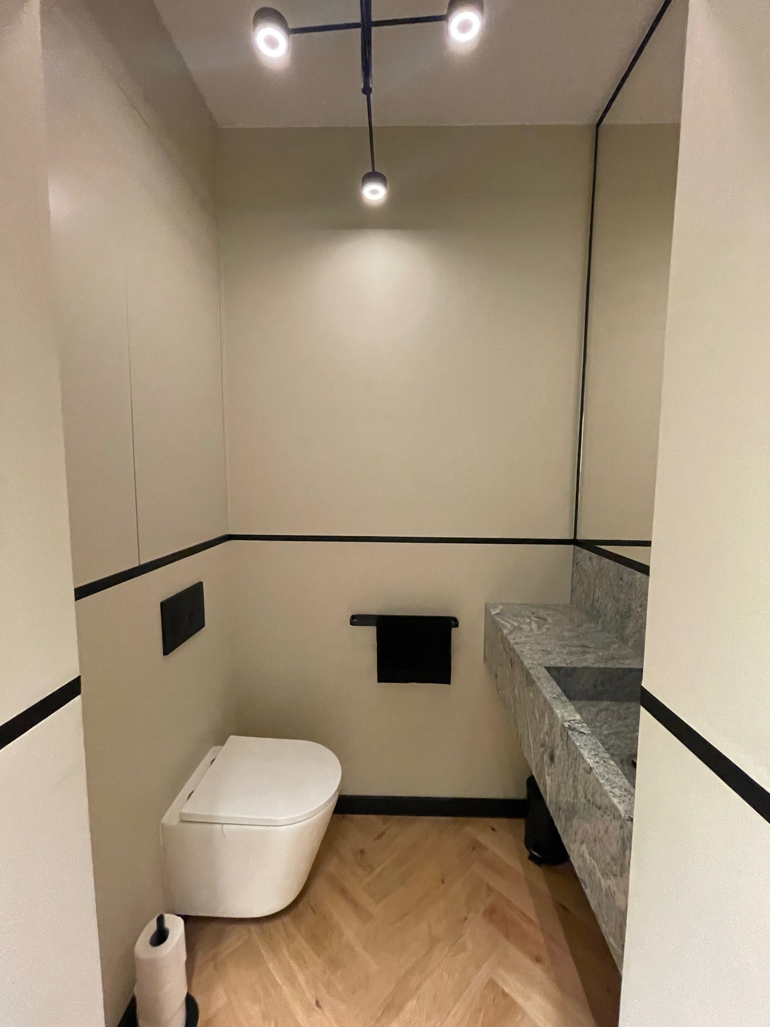 Nowoczesna łazienka z podwieszaną toaletą, umywalką z szarego kamienia i oświetleniem punktowym na czarnym stelażu, wykończona listwami w kolorze czarnym.