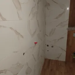 Ściana łazienki wyłożona białymi płytkami imitującymi marmur z brązowymi żyłkami, widoczne otwory na armaturę i fragment drewnopodobnej podłogi.