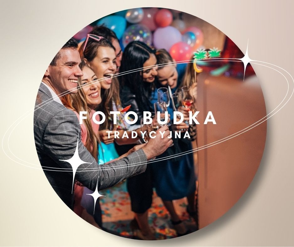 Grupa roześmianych osób, trzymających kieliszki z napojami, pozuje do zdjęcia w tradycyjnej fotobudce z balonami w tle, widoczny fragment męskiej marynarki w kratę.