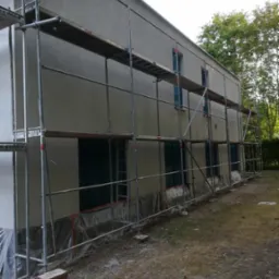 Świeżo otynkowana elewacja domu jednorodzinnego, zabezpieczona folią u podstawy, z rozstawionym rusztowaniem metalowym.