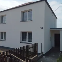 Dwupiętrowy dom z jasną, gładką elewacją, numerem 30 na ścianie i brązowym ogrodzeniem w pierwszym planie, widoczne okna z firankami i zadaszone wejście.
