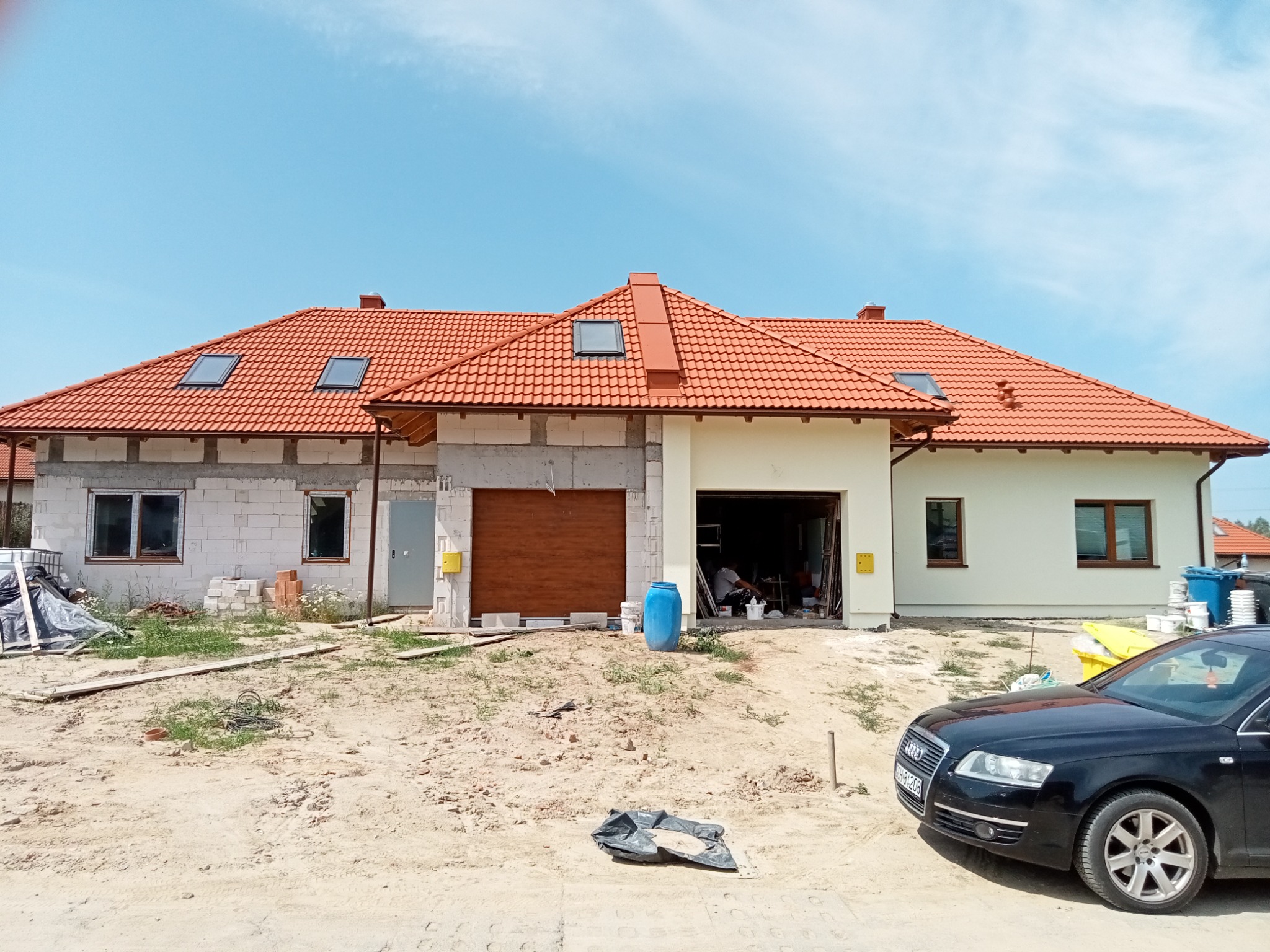 Dom w trakcie budowy lub remontu elewacji, widoczne fragmenty surowego muru, ocieplenia i pomalowanej ściany, na podwórku piasek i materiały budowlane, w tle niebieskie niebo.